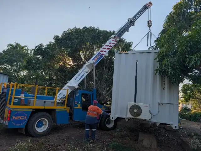 Crane lifting a container building into place between mango trees