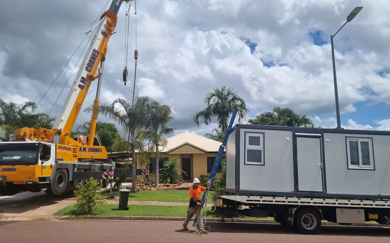 A yellow mobile crane set up in the street, its hook lowered towards the unit on the truck, a rigger in hi-vis carrying a ladder